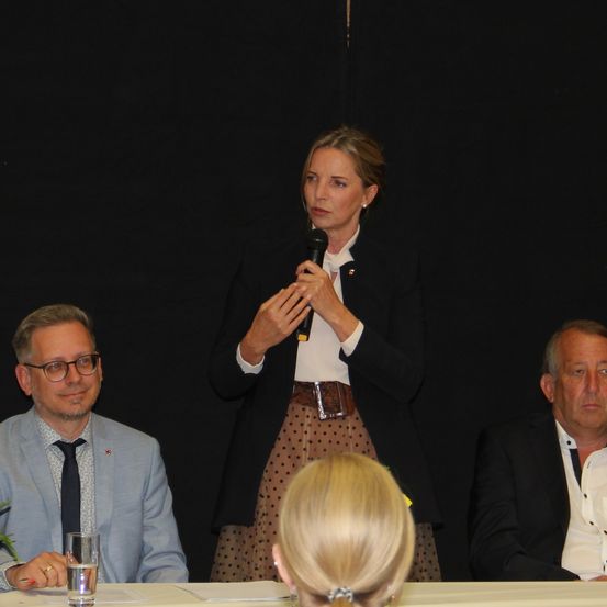 Eine Frau spricht an einem Podium mit einem Mikrofon. Hinter ihr sitzt ein Mann mit Brille und Krawatte am Tisch. Rechts von der Frau sitzt ein weiterer Mann mit kahlem Kopf.