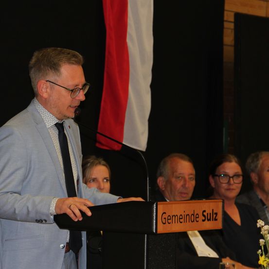 Ein Mann mit Brille und Ring spricht an einem Podium mit Mikrofon. Das Schild darauf lautet 'Gemeinde Sulz'. Hinter ihm sitzen Personen, einige mit Brille, und eine Flagge ist teilweise sichtbar.