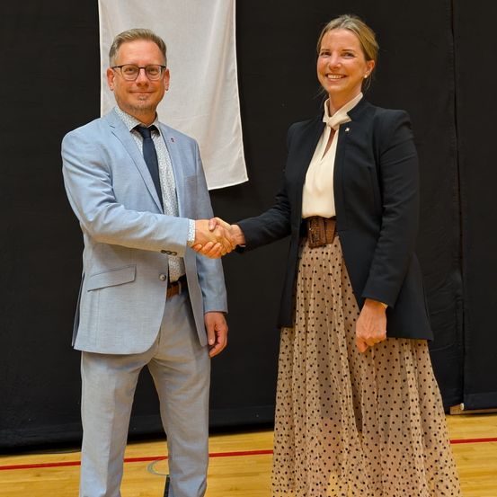 Ein Mann und eine Frau, beide in formeller Kleidung, schütteln sich die Hände vor einem Hintergrund.