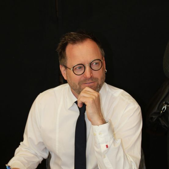 Ein Mann in einem weißen Hemd und einer schwarzen Krawatte sitzt vor einem schwarzen Hintergrund und schaut mit einem Lächeln zur Seite.