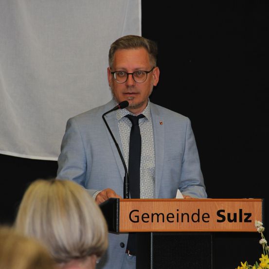 Ein Mann in Anzug und Brille steht hinter einem Podium mit Mikrofon und einem Schild, das Gemeinde Sulz steht.