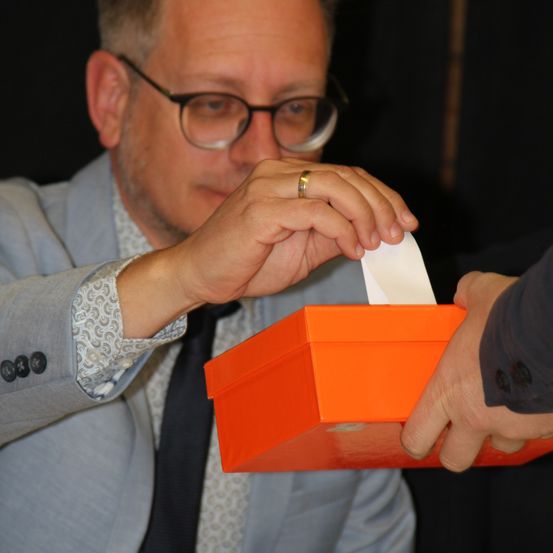 Ein Mann mit Brille und Anzug hält ein Stück Papier und legt es in eine orangefarbene Box.