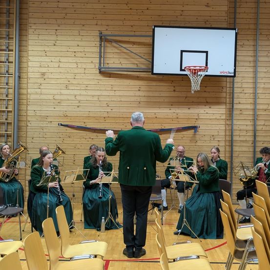 Eine Gruppe von Menschen in grünen Kleidern spielt verschiedene Instrumente in einer Turnhalle mit Holzwänden und einem Basketballkorb.