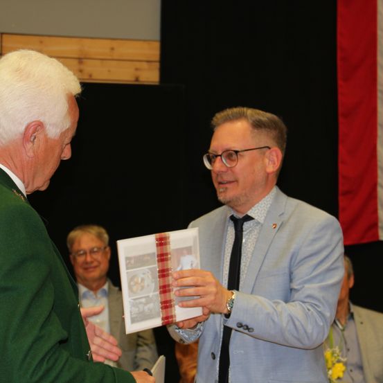 Ein älterer Mann erhält während einer Veranstaltung ein gerahmtes Foto von einem anderen Mann. Hinter ihnen hängt eine Flagge an der Wand.