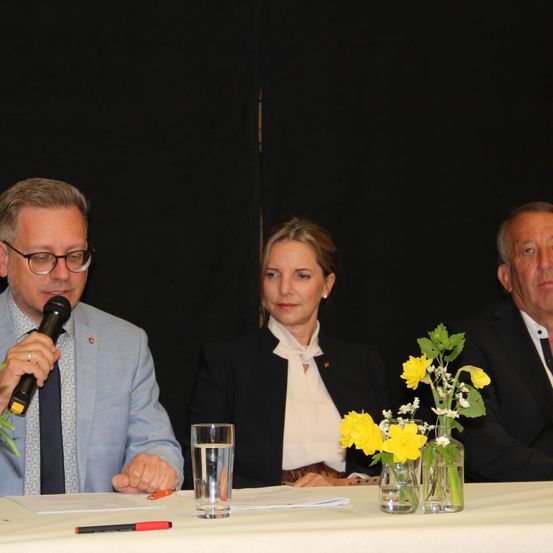 Drei Personen sitzen an einem Tisch und halten wahrscheinlich eine Rede. Ein Mann links hält ein Mikrofon. Eine Frau rechts schaut den Mann an. Vor ihnen steht ein Tisch mit einem Glas Wasser und einer Vase mit gelben Blumen.