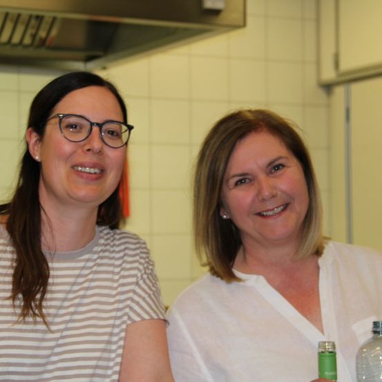 Zwei Frauen lächeln in einer Küche. Die Frau auf der linken Seite trägt eine Brille und ein gestreiftes Shirt. Die Frau auf der rechten Seite hat kurzes Haar und trägt ein weißes Hemd.