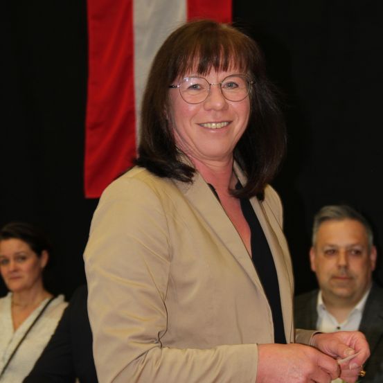 Eine Frau mit Brille lächelt und hält ein Stück Papier in der Hand. Hinter ihr steht eine weitere Frau in einer weißen Bluse, die sie ansieht. Daneben steht ein Mann in einem Anzug mit ernster Miene.