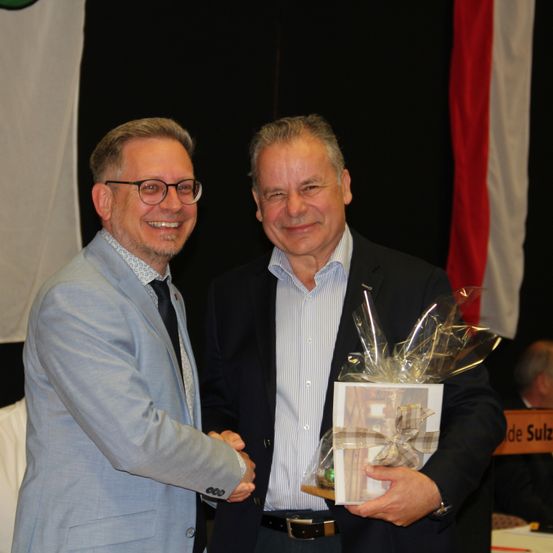 Zwei Männer lächeln und schütteln sich die Hände, einer hält ein in Plastik mit einem Band verpacktes Geschenk. Der Mann links trägt einen hellblauen Anzug mit einem weißen Hemd und einer Brille. Der Mann rechts trägt einen dunklen Anzug und hält das Geschenk. Dahinter steht eine Flagge mit roten und weißen Streifen.