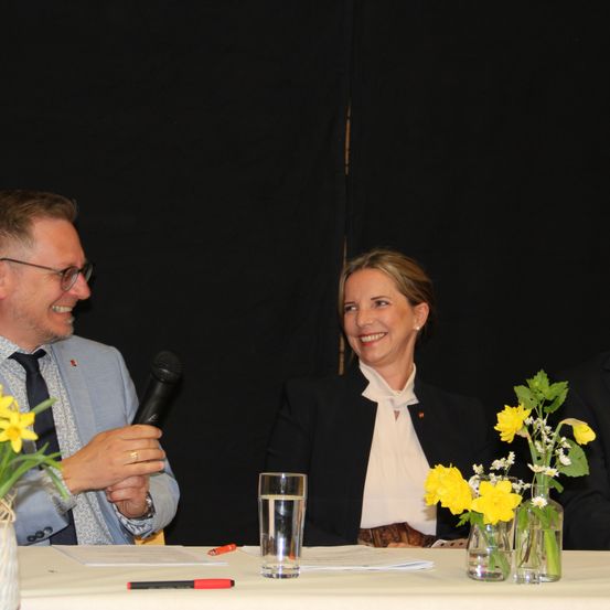 Zwei Personen sitzen bei einer Konferenz an einem Tisch, lächeln und halten ein Mikrofon.