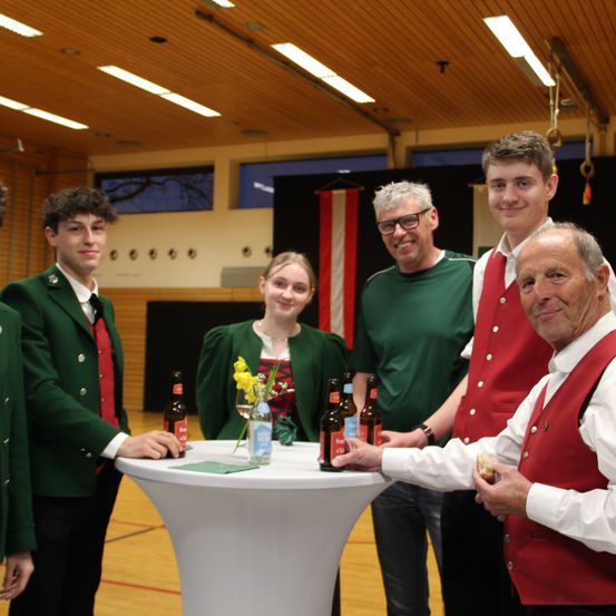 Fünf Personen stehen um einen Tisch mit Flaschen und Essen. Drei von ihnen tragen grün-rote Westen, einer trägt eine Brille und der andere hält Essen.
