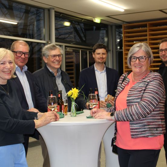 Fünf Personen lächeln und posieren für ein Foto bei einer Veranstaltung. Eine Frau hält ein Weinglas, während ein Mann neben ihr eine Bierflasche hält.