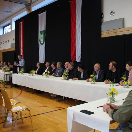 Eine Gruppe von Menschen sitzt an einem langen Tisch mit gelben Blumen in Vasen, ein Mann steht vor ihnen und hält wahrscheinlich eine Rede.