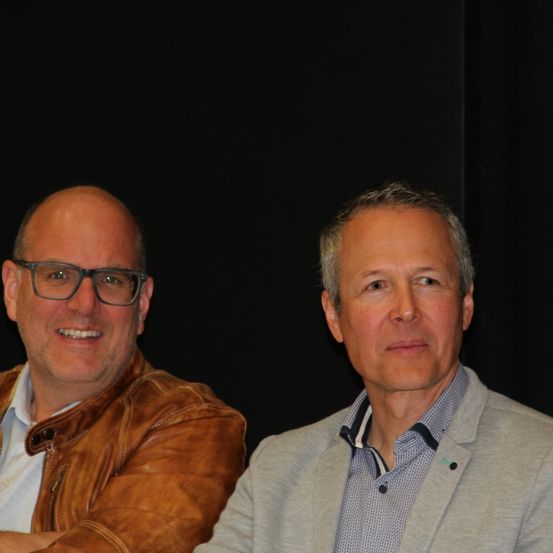 Zwei Männer sitzen zusammen, lächeln und posieren für ein Foto. Ein Mann trägt eine braune Lederjacke und eine Brille. Der andere Mann trägt einen grauen Anzug.