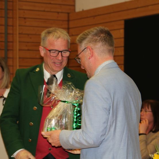Ein Mann in einer grünen Jacke mit Knöpfen überreicht einem anderen Mann in einem blauen Anzug einen Geschenkkorb. Eine Frau mit Brille sitzt hinter ihnen.