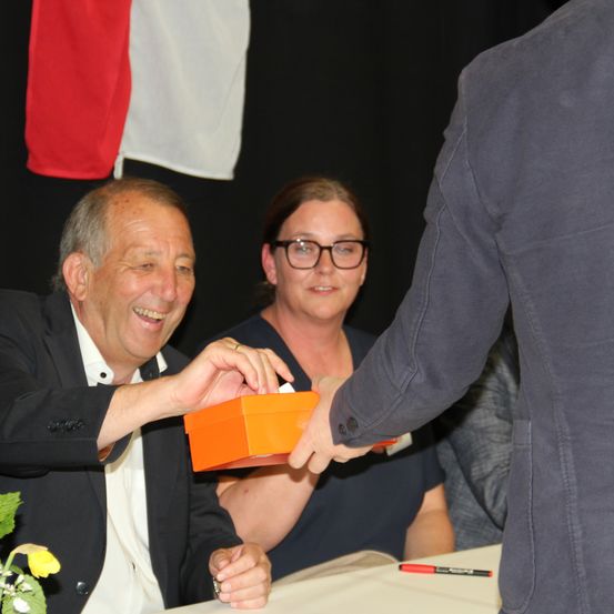 Ein älterer Mann in einem Anzug lächelt, während er eine orangefarbene Box hält und sie einer Frau in einem blauen Shirt reicht.