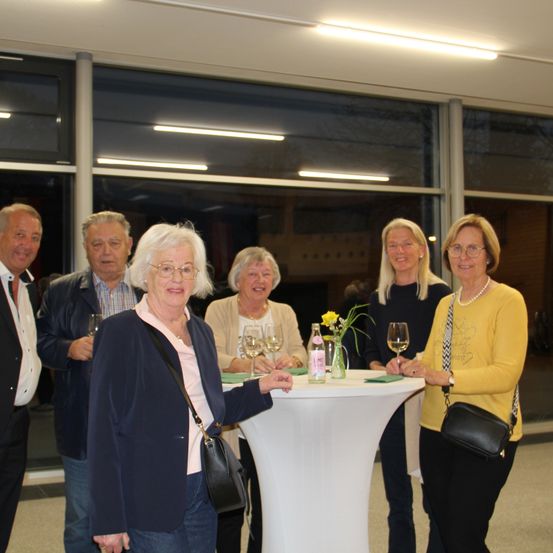 Sechs Personen stehen um einen Tisch in einem modernen Gebäude. Eine Frau hält ein Weinglas.