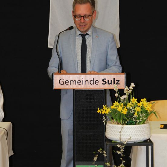 Ein Mann steht hinter einem Podium und hält eine Rede. Das Podium hat ein Schild mit der Aufschrift 'Gemeinde Sulz'.