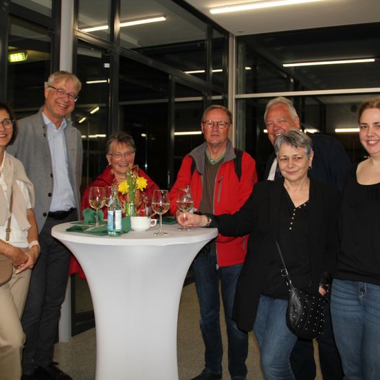 Eine Gruppe lächelnder Menschen steht um einen Tisch mit Weinläsern, einer Vase mit gelben Blumen und einer Flasche herum, mit einer Glaswand dahinter.