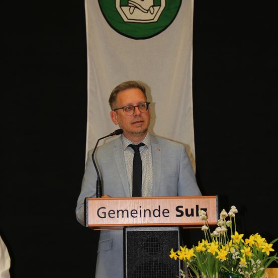 Ein Mann steht an einem Podium mit einem Mikrofon vor einem Banner, das Gemeinde Sulz steht.