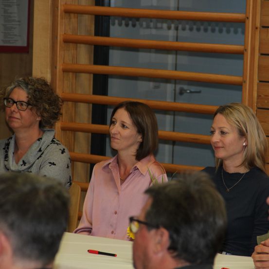 Drei Frauen und zwei Männer sitzen an einem Tisch bei einer Besprechung. Die Frauen tragen Brillen und Halsketten.