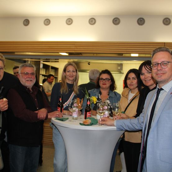 Mehrere Personen, darunter ein Mann in Anzug, stehen um einen runden Tisch in einer Inneneinrichtung. Sie halten Gläser mit Wein und haben Blumen und Essen auf dem Tisch. Der Raum ist gut beleuchtet.