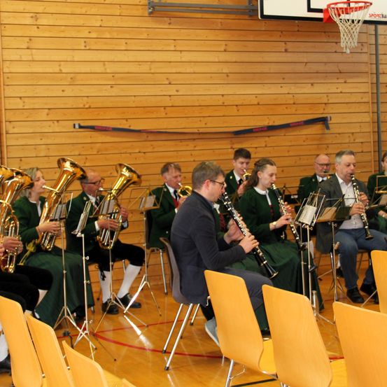 Eine Gruppe von Musikern in grüner Kleidung spielt in einer Turnhalle. Sie sitzen auf Stühlen mit Notenständern vor ihnen. Ein Basketballkorb ist an der Wand montiert.