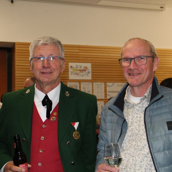 Zwei Männer lächeln für die Kamera. Der Mann auf der linken Seite hält eine Flasche Bier und der Mann auf der rechten Seite hält ein Glas Wein.