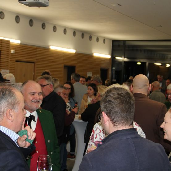 Mehrere Personen sind in einem Raum versammelt. Ein Mann in einem grünen Vest spricht mit einem anderen Mann, und eine Frau in einem Blumenhemd spricht mit einem Mann in einem braunen Jackett. Ein Projektor ist an der Decke montiert, und eine Tafel ist an der Wand befestigt.