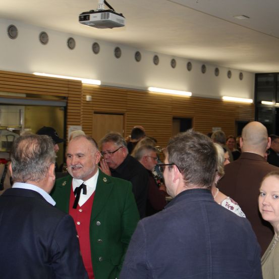 Eine Gruppe von Menschen steht in einem Raum. Ein Mann in der Mitte lächelt und spricht mit anderen. Er trägt eine grüne Weste mit einem roten Westchen und einer schwarzen Schleife. Der Raum hat eine Projektor und Lampen an der Decke. Es gibt Holzwände und eine Tür an der Seite. Einige Leute stehen in der Nähe der Tür. Es gibt eine verschwommene Ansicht des Raumes.