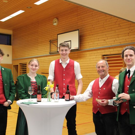 Fünf Personen stehen in einem Fitnessstudio um einen Tisch herum, lächeln und posieren für ein Foto. Der Mann in der Mitte hält eine Flasche, während die anderen Weingläser und Blumen auf dem Tisch haben.