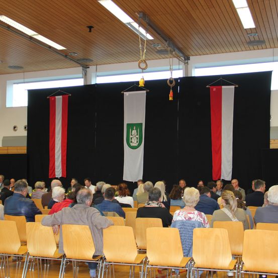 Ein Saal mit Holzstühlen, in dem Menschen in Reihen sitzen. Es gibt eine Bühne mit schwarzem Hintergrund und einer Flagge mit grünem Emblem sowie zwei rot-weißen Flaggen.