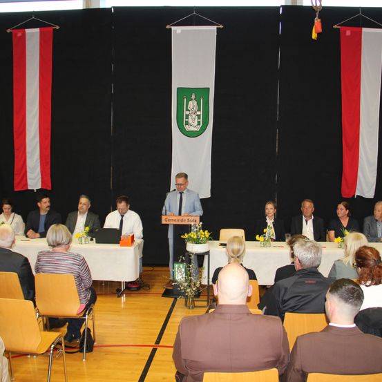 Eine Gruppe von Menschen sitzt um einen Tisch herum und hört einem Mann zu, der an einem Podium spricht. Das Podium hat ein Schild mit der Aufschrift 'Gemeinde Sulz'. Der Tisch hat eine gelbe Blumendekoration. Hinter dem Mann hängt ein weißer Banner mit einem grünen Schild.