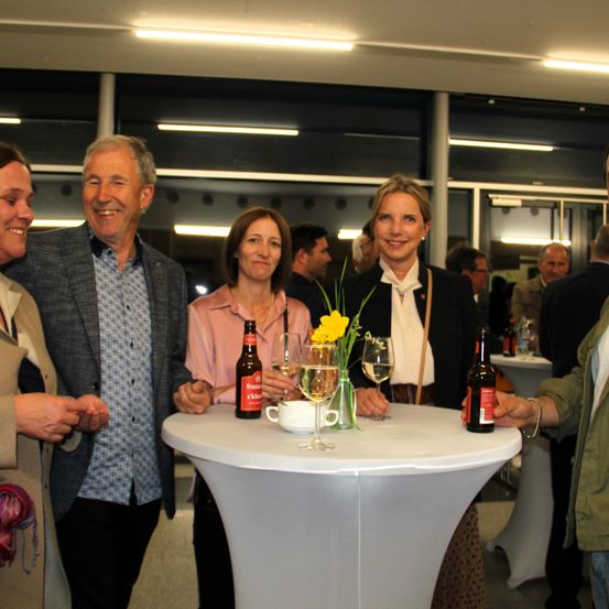 Eine Gruppe von Menschen steht um einen Tisch herum, auf dem sich eine Blumenarrangement, Flaschen und Weingläser befinden.