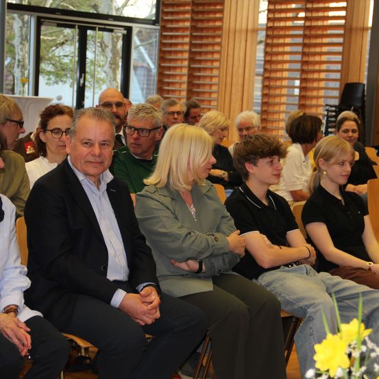 Eine Gruppe von Menschen sitzt in einem Konferenzraum, mit einem Mann und einer Frau mit Brille und einer Frau mit blonden Haaren und einer Uhr.