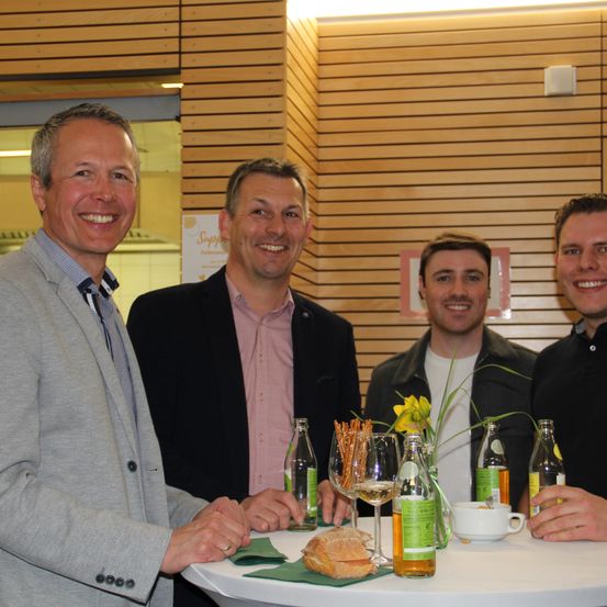 Vier Männer lächeln in die Kamera. Der Mann auf der linken Seite trägt einen grauen Anzug. Der Mann neben ihm trägt einen schwarzen Mantel und ein rosa Hemd. Der Mann auf der rechten Seite trägt einen schwarzen Mantel. Ein Tisch mit Brot, Weingläsern und Flaschen steht vor ihnen.