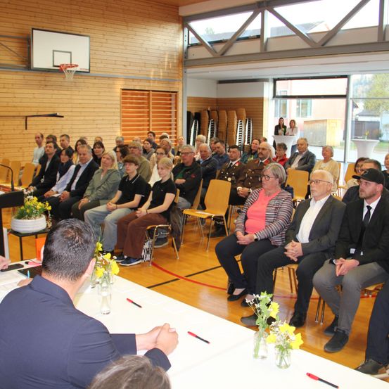 Eine Gruppe von Menschen sitzt in einer Turnhalle und hört einem Sprecher vorne zu. Der Raum hat Holzwände, Glasfenster und einen Basketballkorb. Es gibt Blumen auf den Tischen und Stühlen.