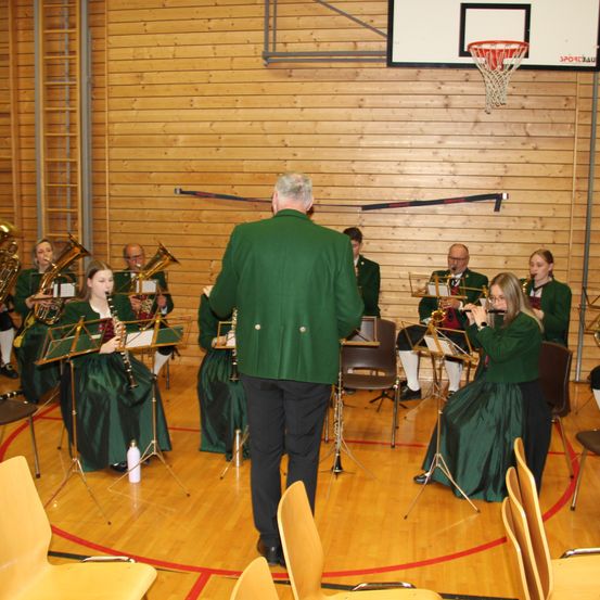 Ein Bläserensemble spielt in einer Turnhalle. Mehrere Musiker spielen verschiedene Instrumente, darunter Trompeten, Posaunen und Flöten. Die Band steht auf einem Holzboden, und ein Basketballkorb ist an der Wand zu sehen.
