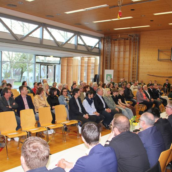 Ein Saal mit Holzwänden und einer Glastür, in dem eine Gruppe von Menschen in Stuhlreihen sitzt. Ein Banner ist an der Wand montiert und eine Leiter steht rechts. Einige Leute stehen in der Nähe der Glastür.