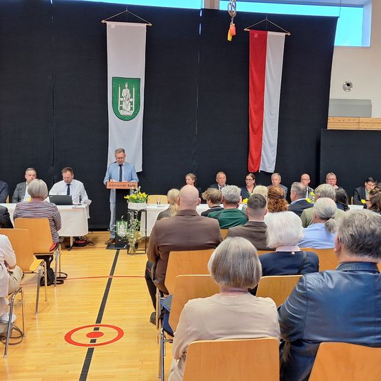 Eine Gruppe von Menschen sitzt in Stühlen in einem Saal mit einem Podium. Ein Mann steht am Podium und spricht die Zuhörer an. Eine rot-weiße Flagge hängt an der Wand.