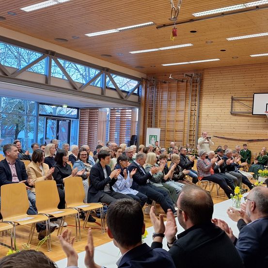 Eine Gruppe von Menschen versammelt sich in einem Raum mit Holzdecke, großen Fenstern und einem Basketballkorb. Einige Leute klatschen, während andere sitzen. Ein Mann steht und klatscht vor einer Gruppe von Menschen. Eine gelbe Blume steht in einer Vase auf einem langen Tisch.