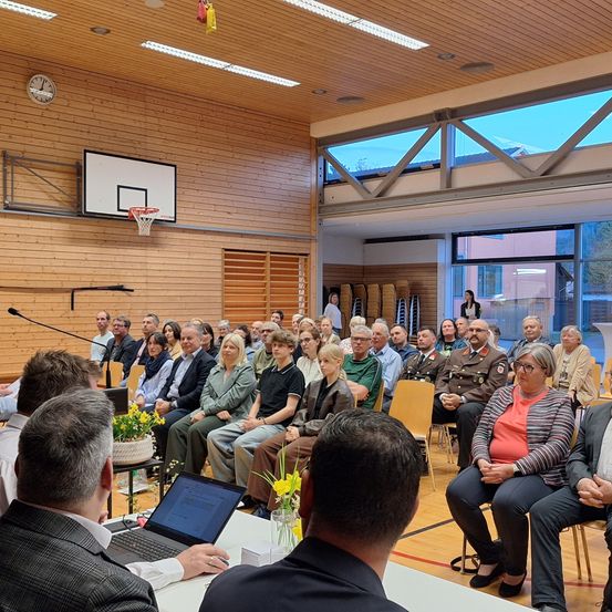 Eine Gruppe von Menschen sitzt in einer Halle mit Holzwänden, einem Basketballkorb und einer Uhr an der Wand. Eine Person vorne hat einen Laptop auf dem Tisch.