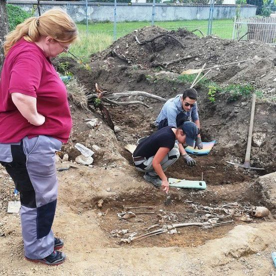 Drei Personen arbeiten in einer Grube, möglicherweise bei einer archäologischen Ausgrabung. Eine Person kniet und untersucht etwas, während eine andere in der Nähe steht.