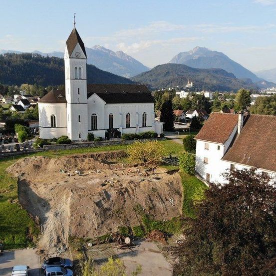 Luftaufnahme einer Kirche mit einem Kirchturm, umgeben von einem Friedhof und einem Bergrücken, mit einem Gebäude rechts und einigen geparkten Autos im Vordergrund.