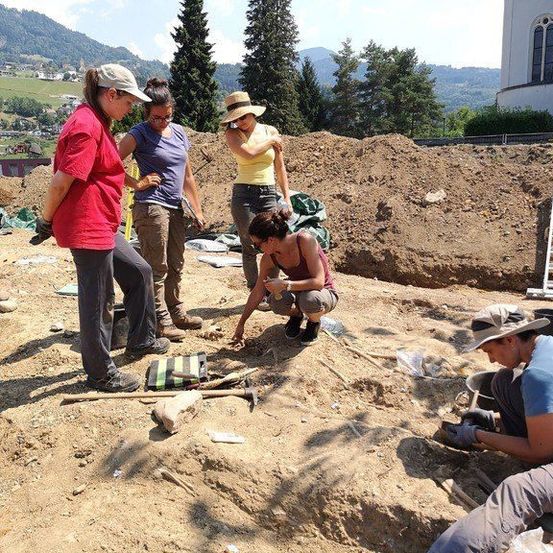 Archäologen untersuchen eine Stelle, einer kniet, um etwas zu inspizieren. Verschiedene Werkzeuge und Gegenstände sind um sie herum verstreut. Die Umgebung ist im Freien, mit einer Kirche und einer bergigen Landschaft im Hintergrund.