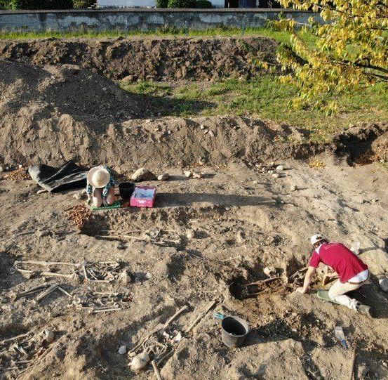 Archäologen graben an einer Stelle mit sichtbaren Knochen und Werkzeugen, wobei eine Person sitzt und zwei andere knien.