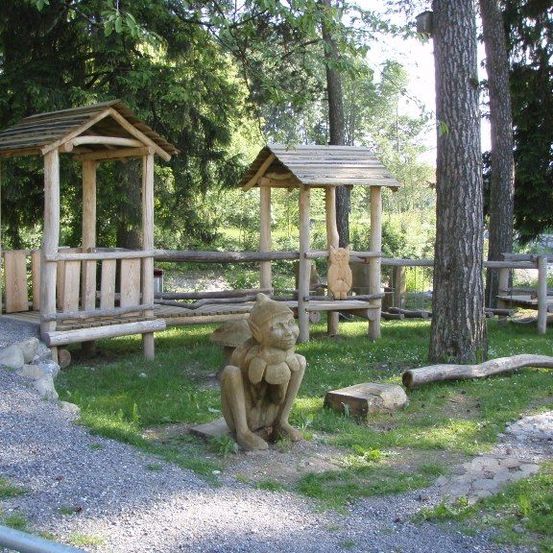 Ein hölzerner Spielbereich mit einer kleinen Holzhütte und einer geschnitzten Holzfigur eines Gnoms auf dem Rasen in einem Wald.