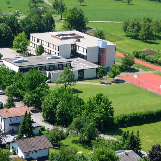 Ein Luftbild eines Schulkomplexes mit einem großen Gebäude, kleineren angeschlossenen Gebäuden und einer roten Laufbahn. Bäume und grüne Felder umgeben das Gebiet, mit einem klaren Himmel darüber.