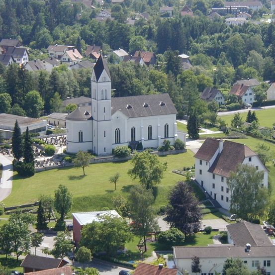 Luftaufnahme eines Dorfes mit einer großen weißen Kirche und einem braun gedeckten Haus im Vordergrund. Das Dorf ist von grünen Bäumen und Gras umgeben.