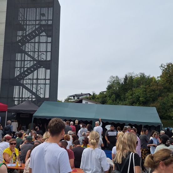 Eine Menschenmenge versammelt sich im Freien unter einem grünen Zelt, wobei ein großes Gebäude mit einer Feuertreppe im Hintergrund zu sehen ist. Bäume und Häuser sind in der Ferne erkennbar.