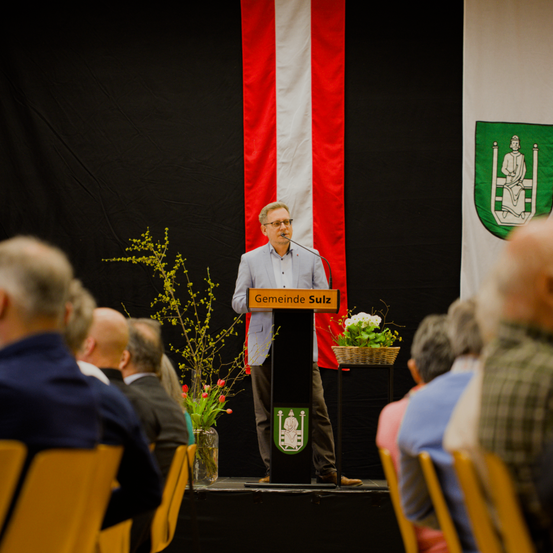 Ein Mann steht an einem Podium auf der Bühne und spricht ein Publikum an. Hinter ihm befindet sich eine rot-weiße Flagge mit einem grünen Wappen. Blumen und Pflanzen schmücken die Bühne.